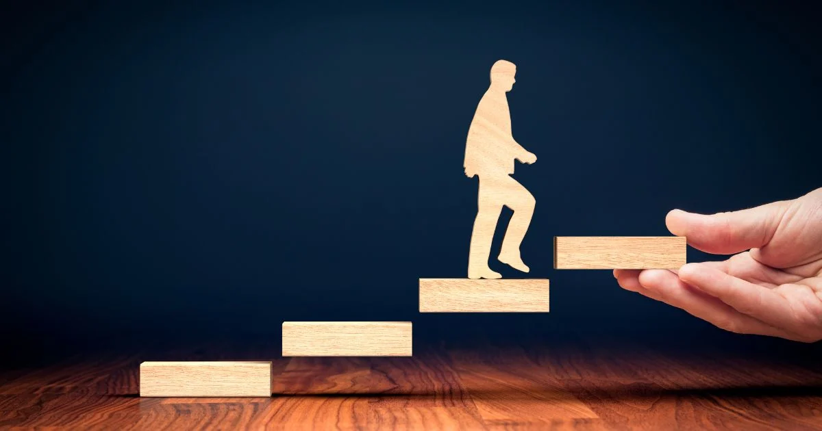 A wooden silhuete of a man walking up wooden blocks against the dark blue background. From the right side a hand is holding up the next block
