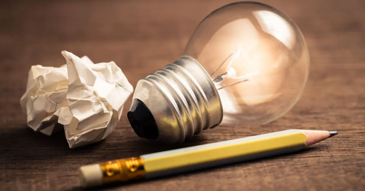 A crumpled piece of white paper, a lightbulb and a pencil on a wooden surface